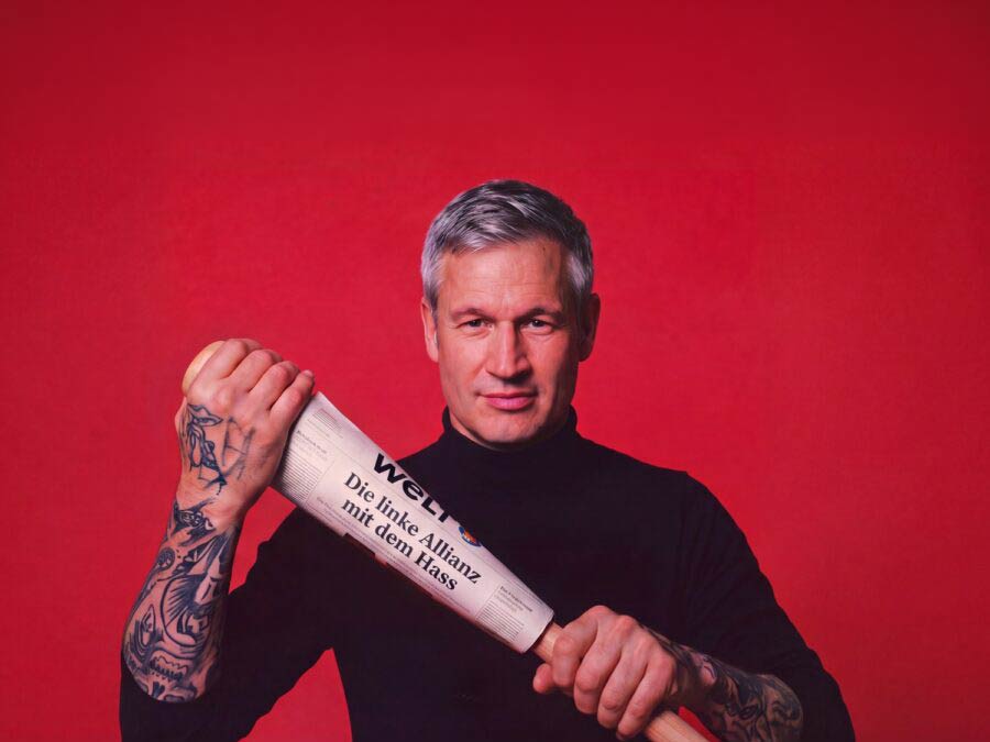 Satirical AI montage: Ulf Poschardt, wearing a black turtleneck sweater with tattooed underarms, holds a baseball bat against a red background, wrapped in a world edition with the headline »The Left Alliance with Hate«